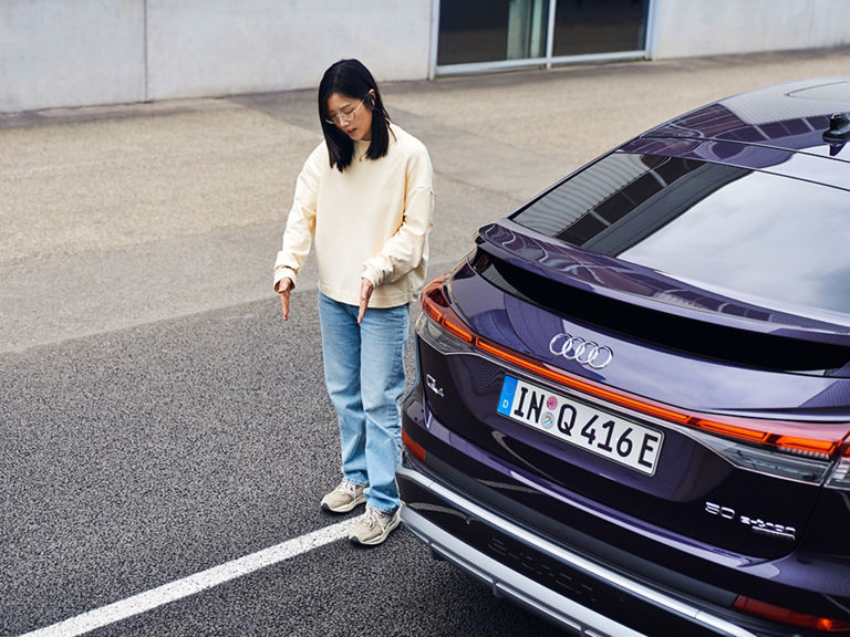 Tiantian erklärt etwas und steht neben einem lilanen Audi
