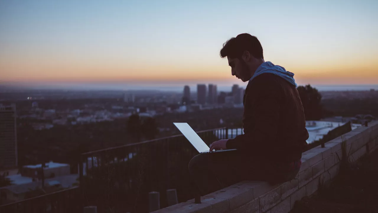 A man sits on a wall at sunrise with his laptop on his lap