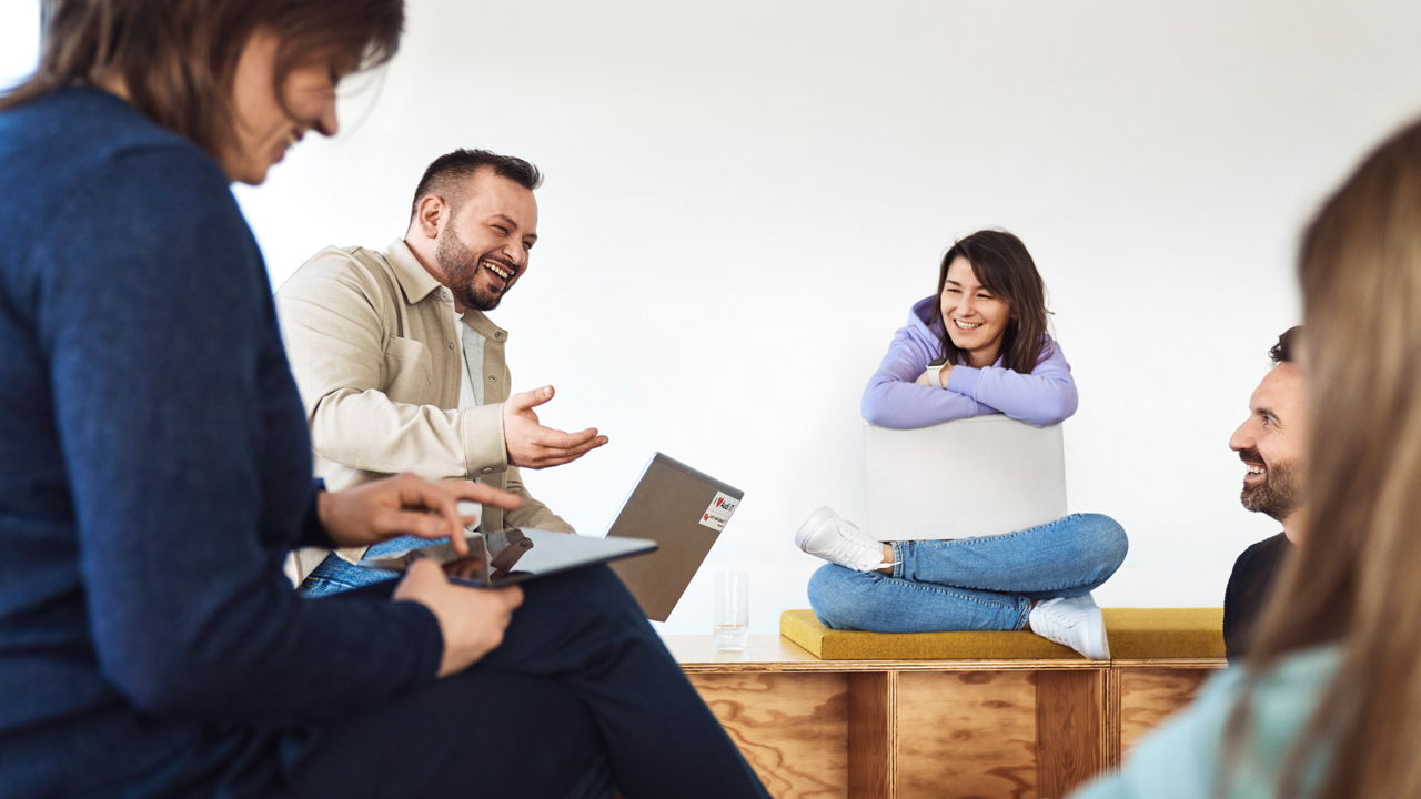 Employees sit in a circle at an informal meeting. A woman sits cross-legged on a wooden bench and leans on her laptop while a colleague talks. 