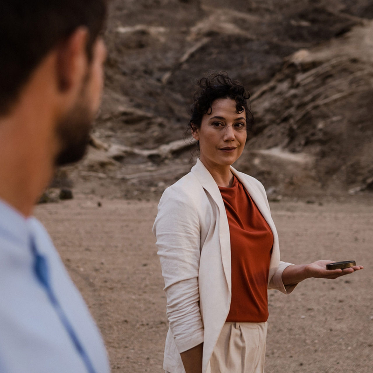 The back of a person can be seen in the foreground. In the centre of the picture is a woman looking in the direction of the other person. Both are in a desert environment.