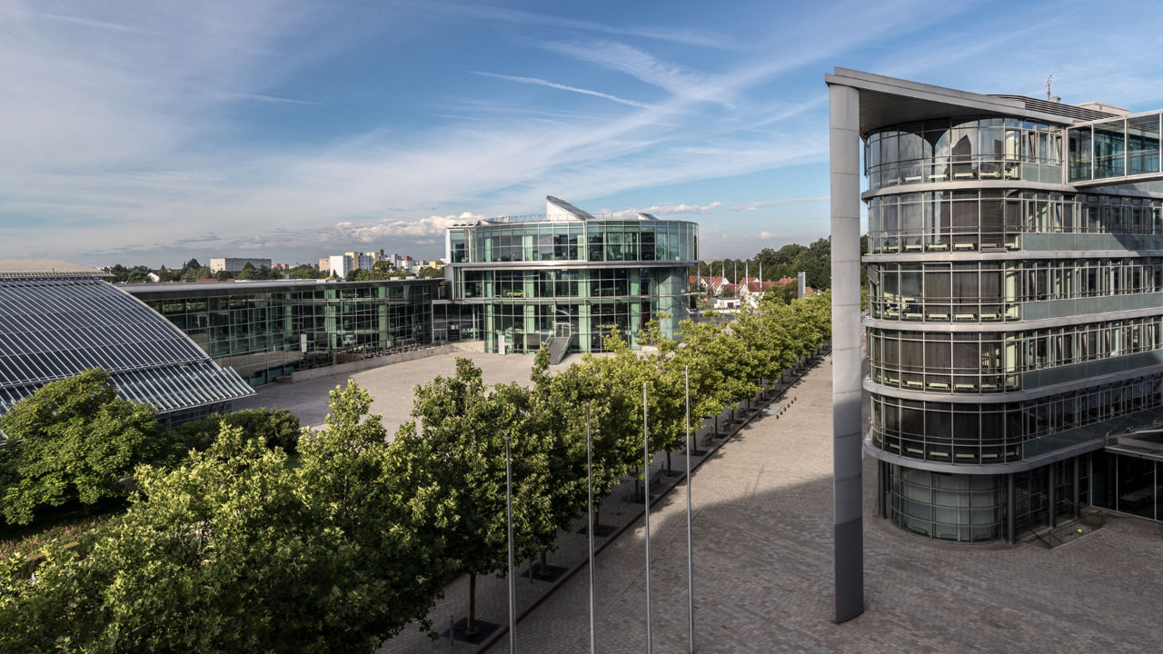 A modern building complex with large glass façades and curved shapes, surrounded by a row of trees and paved paths under a clear blue sky.