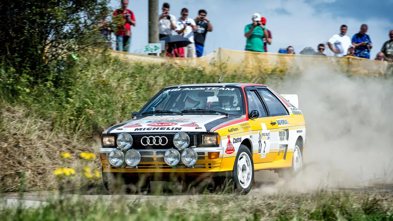 Eine bunt beklebter Audi fährt auf einer Sandstrecke und wirbelt Staub auf. Im Hintergrund stehen Zuschauer, die Fotos machen.