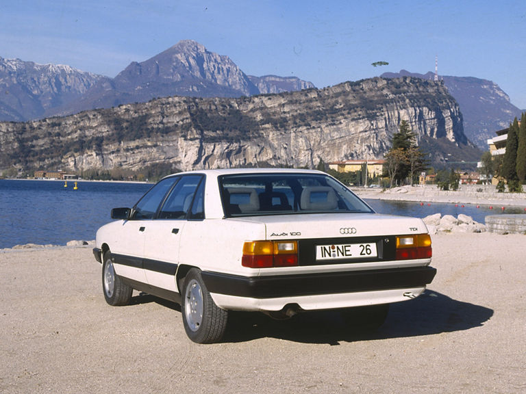 Ein weißer Audi 100 TDI in der Heckansicht vor einer Bergkulisse mit Bergsee