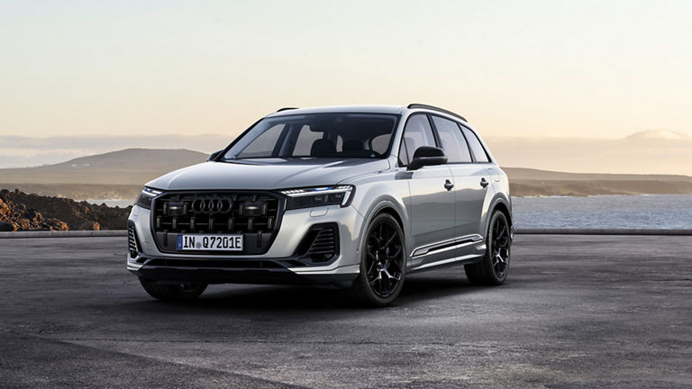 A white Audi Q7 TFSI e with a black radiator grille and black rims stands in front of a picturesque coastal landscape with rolling hills in the background, while the evening sun bathes the sky in warm colors.