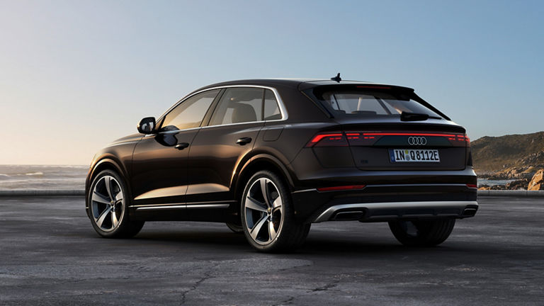 A black Audi Q8 TFSI e stands with its elegant silhouette in front of a picturesque coastal landscape. The setting sun is reflected on the bodywork, while the red LED light strip at the rear stands out strikingly.