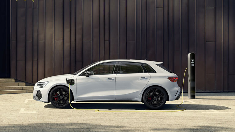 A white Audi A3 Sportback TFSI e stands at the side of a charging station while the yellow charging cable is connected. In the background, a modern dark metal façade with a tree whose leaves protrude into the scene.