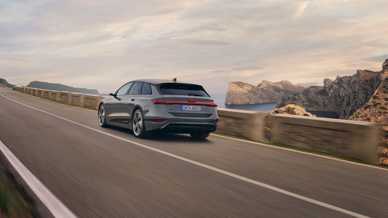 A gray Audi A6 Avant drives along a winding coastal road with an impressive rocky landscape and sea view in the background. The scenery is accompanied by a warm evening atmosphere.
