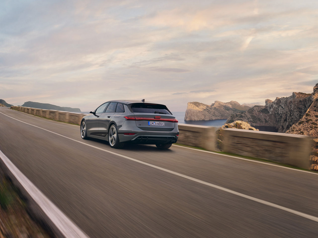 A gray Audi A6 Avant drives along a winding coastal road with an impressive rocky landscape and sea view in the background. The scenery is accompanied by a warm evening atmosphere.