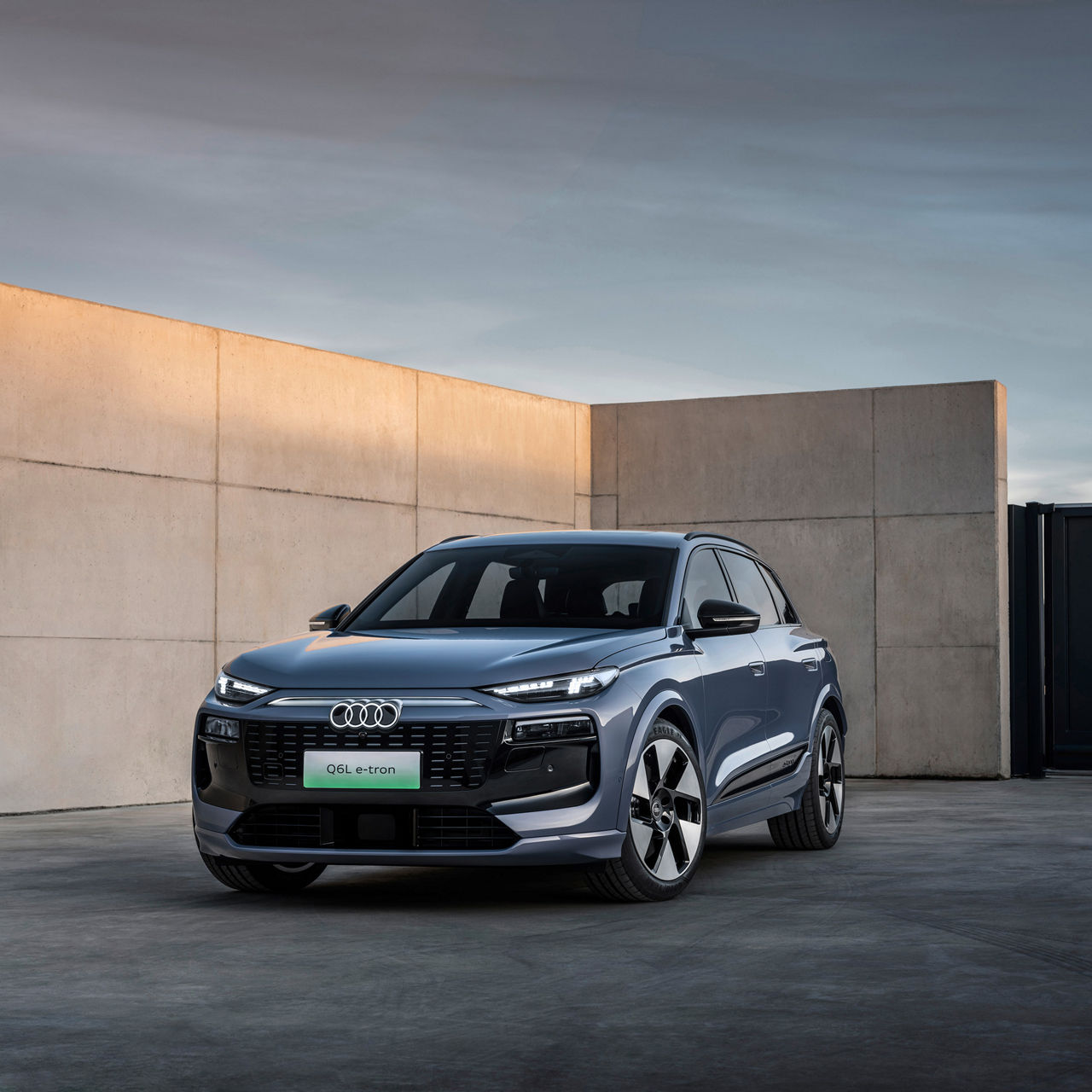 An Audi Q6L e-tron in metallic blue paintwork stands in front of a modern concrete wall and a black gate, while the sky glows in soft evening colors.