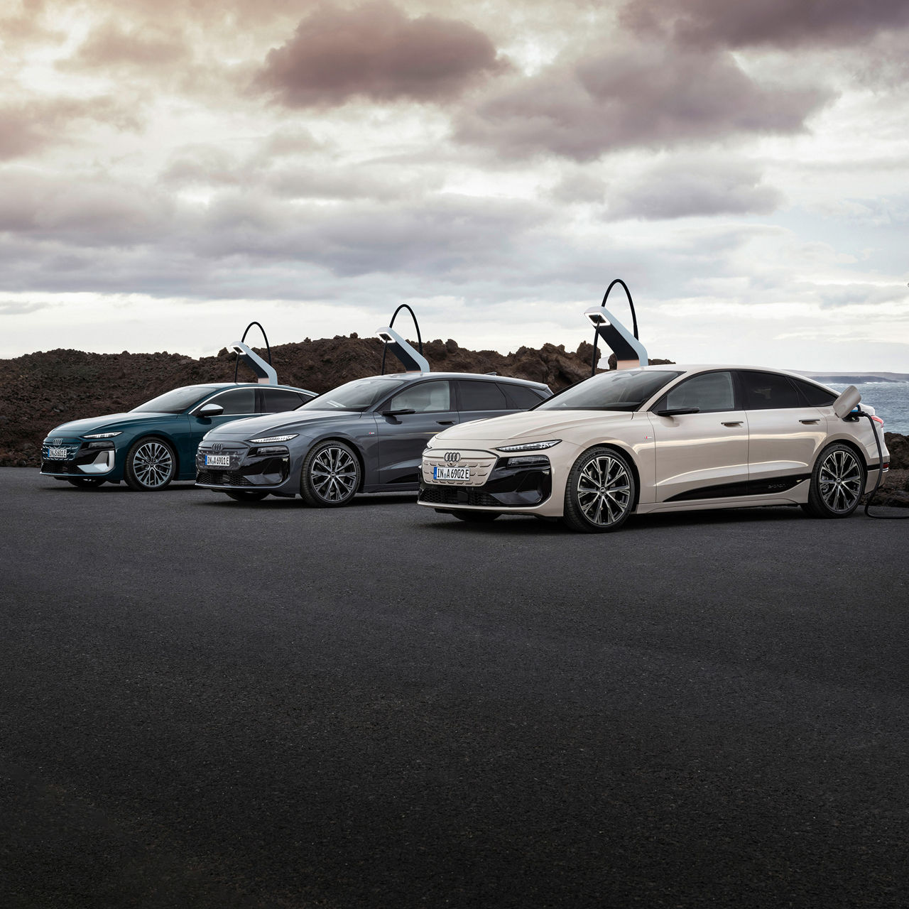 Three Audi Q6 e-tron vehicles in different colors are parked at a charging station in front of a rocky coastal landscape with a cloudy sky. The vehicle in front is connected to the charging station.