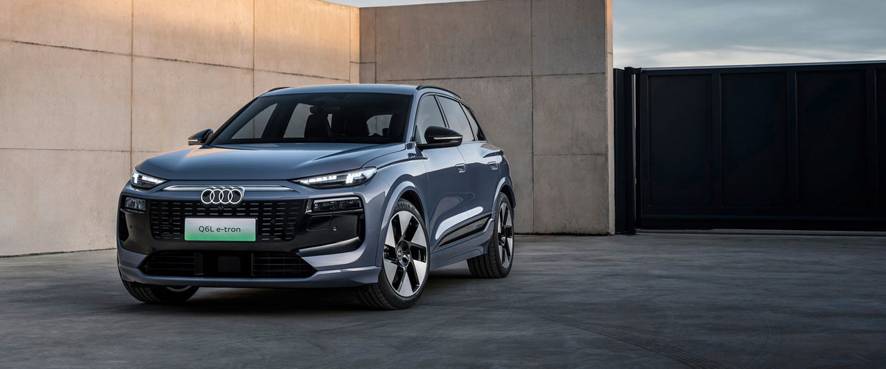 An Audi Q6L e-tron in metallic blue paintwork stands in front of a modern concrete wall and a black gate, while the sky glows in soft evening colors.