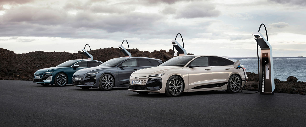 Three Audi Q6 e-tron vehicles in different colors are parked at a charging station in front of a rocky coastal landscape with a cloudy sky. The vehicle in front is connected to the charging station.