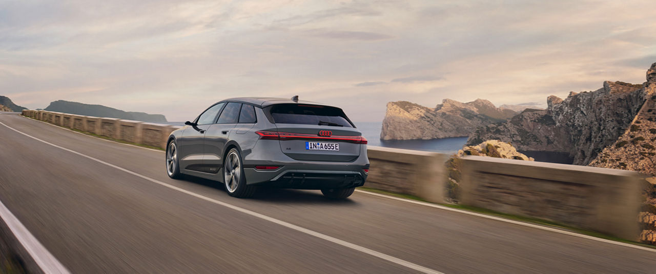 A gray Audi A6 Avant drives along a winding coastal road with an impressive rocky landscape and sea view in the background. The scenery is accompanied by a warm evening atmosphere.
