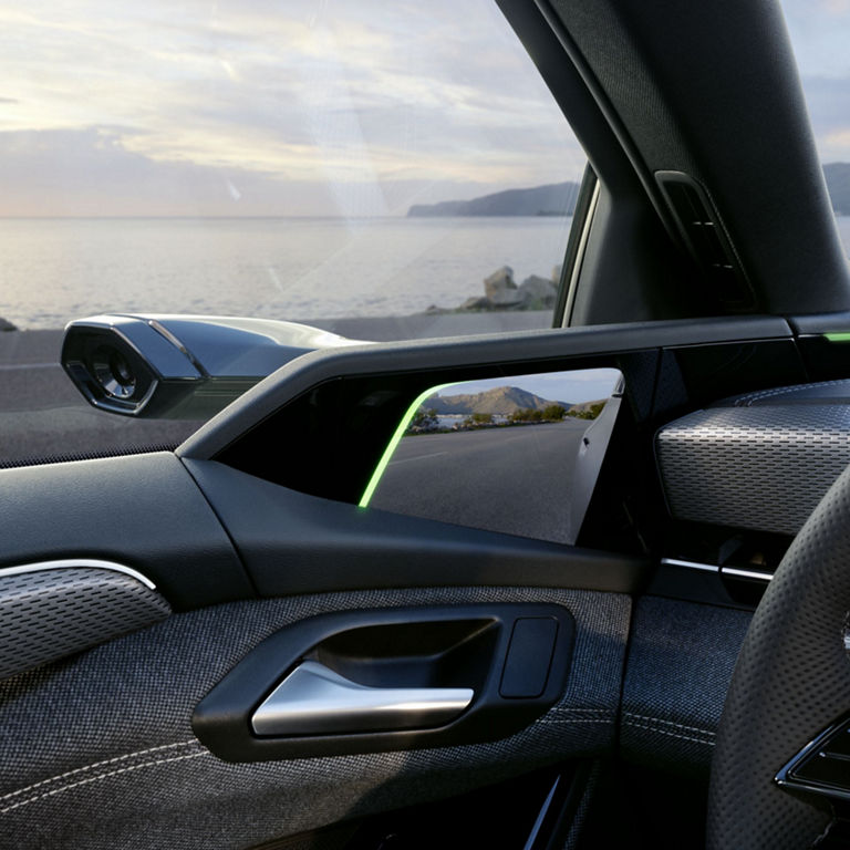 Detailed view of a digital exterior mirror in an Audi. The camera on the outside of the vehicle transmits the image to a screen in the interior. A coastal road with a view of the sea and mountains can be seen in the background.