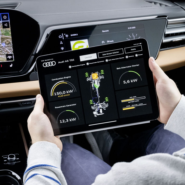 A driver holds a tablet in an Audi A5 TDI. A real-time diagnosis of the vehicle is displayed on the screen, including data on the combustion engine, alternator and battery. The dashboard with a digital display and navigation system can be seen in the background.