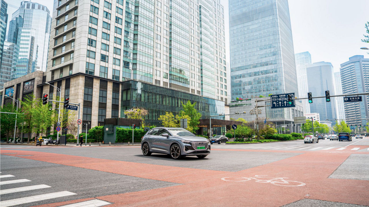 Ein silberner Audi fährt auf einer breiten Straße inmitten einer modernen Stadtlandschaft mit hohen Gebäuden und viel Glasarchitektur.