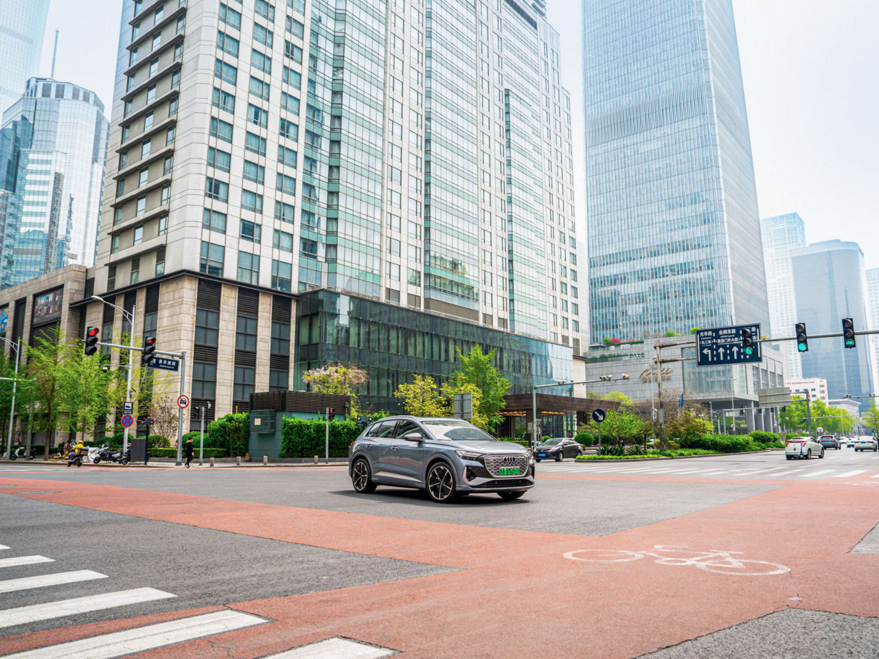 Ein silberner Audi fährt auf einer breiten Straße inmitten einer modernen Stadtlandschaft mit hohen Gebäuden und viel Glasarchitektur.