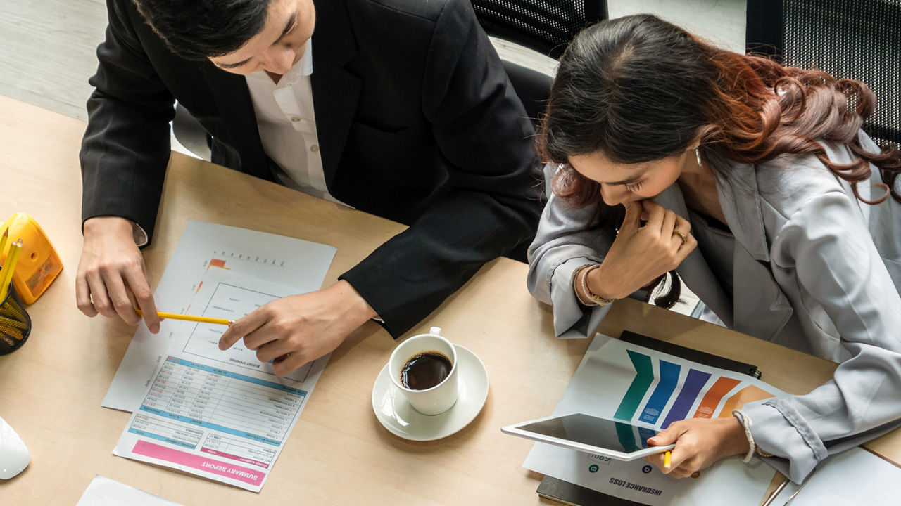 Ein Mann und eine Frau in Business-Kleidung arbeiten zusammen an einem Tisch, der mit Berichten, einem Tablet und einer Kaffeetasse bedeckt ist.