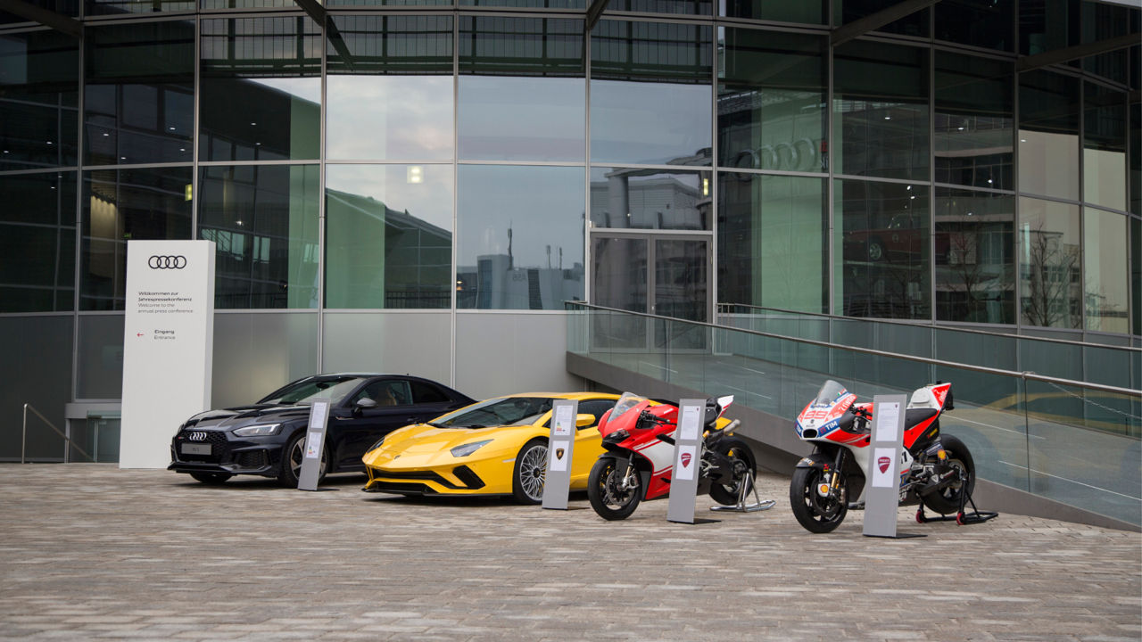  Ein Audi, ein Lamborghini und zwei Ducati Motorräder vor dem Eingang des Audi Forum Ingolstadt