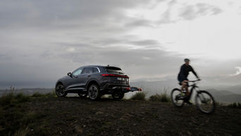 An Audi Q5 is parked on a cliff with a mountain panorama. A cyclist rides away from the car.