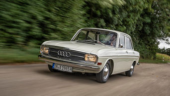 An Audi F103 with an H license plate (Oldtimer) drives along a country road at the edge of a forest.