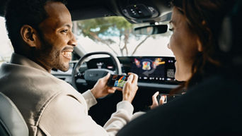 Zwei Personen spielen ein digitales Kartenspiel in einem Audi, angezeigt auf Smartphones und dem zentralen Bildschirm.
