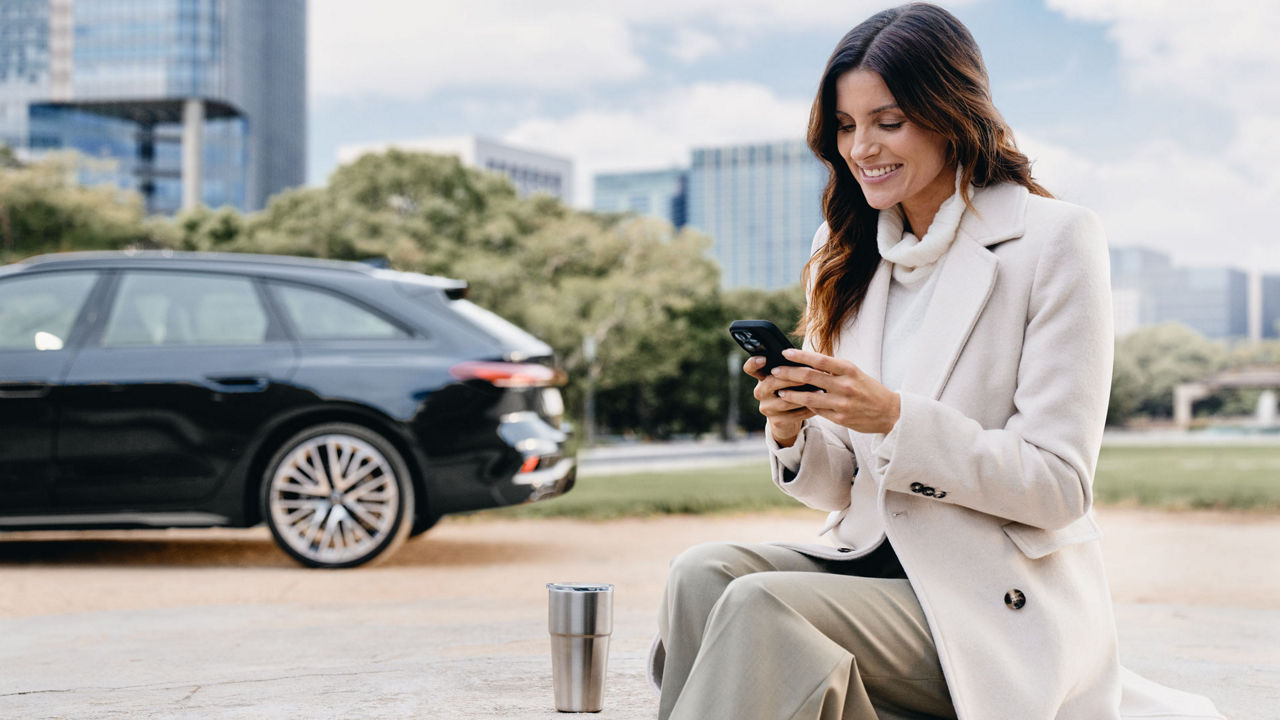 A woman sits on a bench, smiling at her phone screen. In the back is a black Audi parking in front of a tree line.