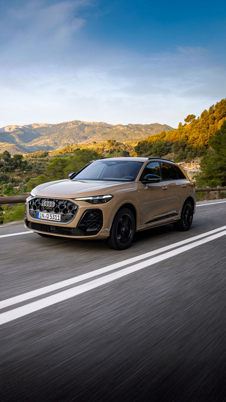 An Audi Q5 SUV drives along a mountain road. In the background, green hills bathed in golden sunlight.