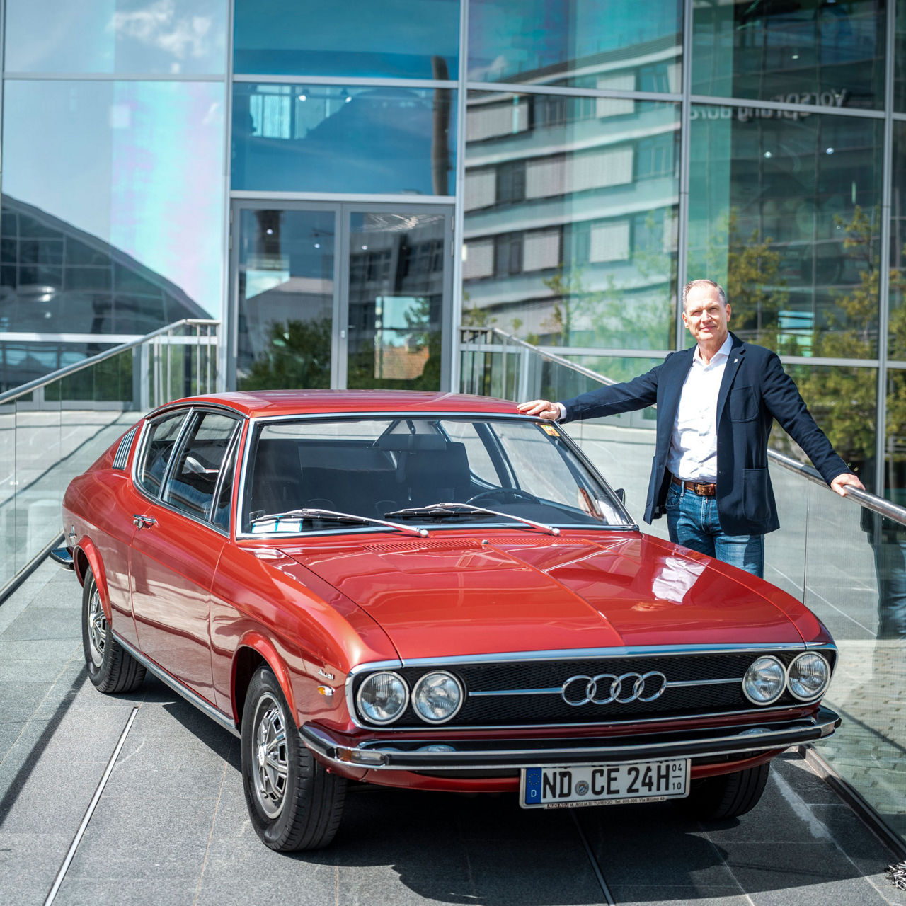 Rene Marc Heinze in a dark jacket, white shirt and jeans leans against his red Audi 100 on the ramp of the Audi museum mobile