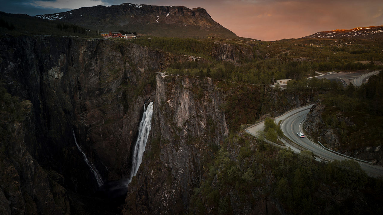 Panoramaansicht der gewaltigen Bergmassive im Westen Norwegens mit Blick auf den Vøringsfossen Wasserfall und kurvenreicher Straße, auf der klein der Audi A6 Sportback e-tron performance zu sehen ist.