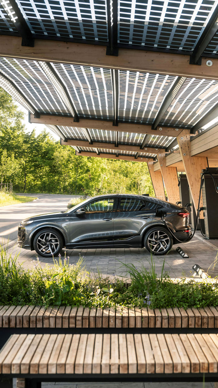 Dunkelgrauer Audi Q6 Sportback e-tron lädt an einer Ladesäule in einer holzüberdachten Ladestation.