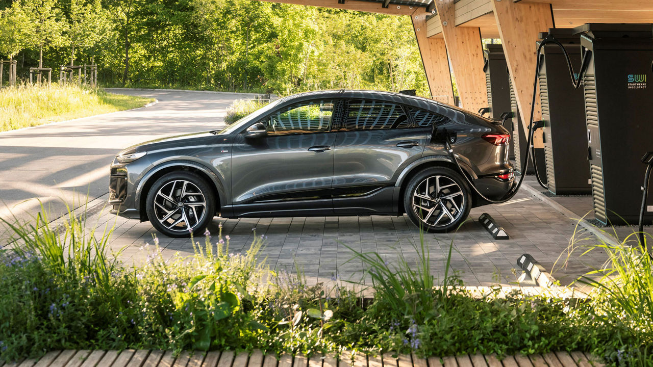 Dark grey Audi Q6 Sportback e-tron charging at a charging station in a wooden-roofed charging station.