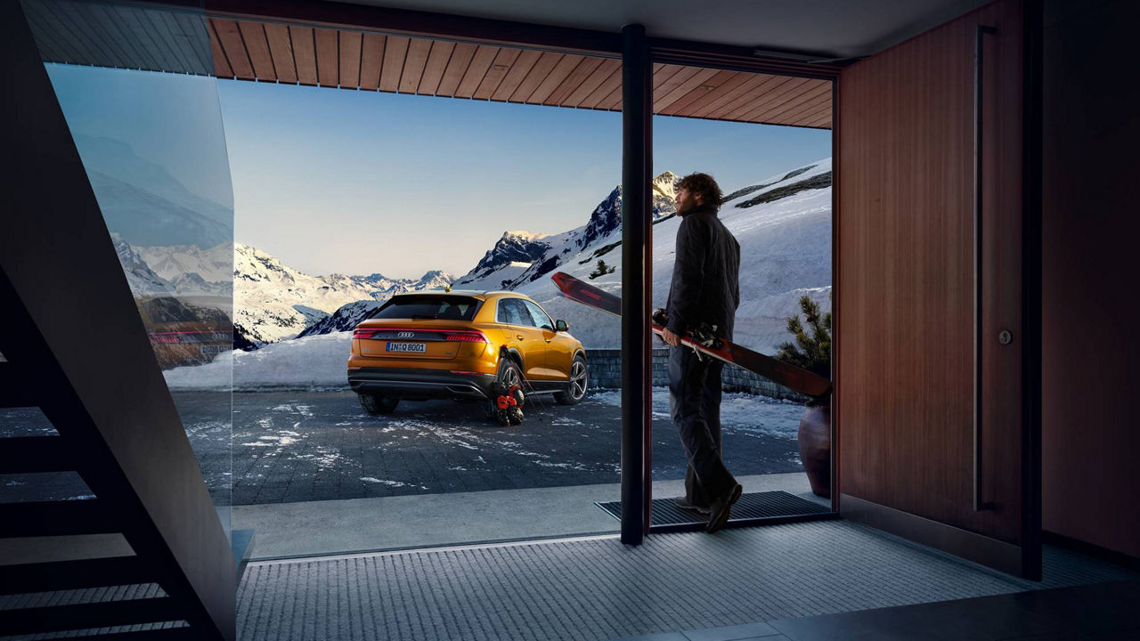 A man looks out of the panoramic window of a modern ski lodge at a snow-covered mountain backdrop and a yellow Audi parked in front of the building.