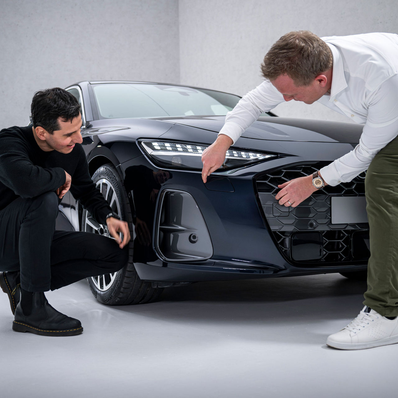 Francesco d’Amore (crouched down) and Sebastian Weiper (bent over) examine the front end of a black Audi A6 Sedan, specifically looking at the side air intakes, the so-called air curtains.