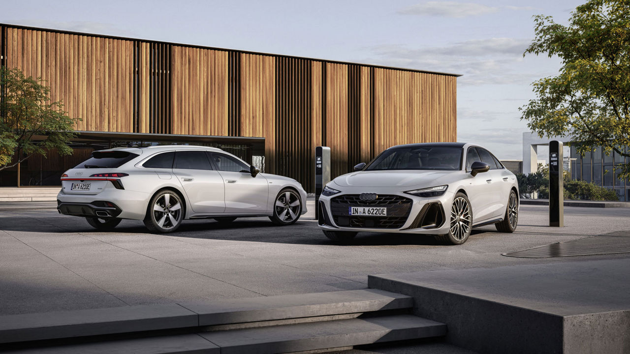 Audi A6 e-hybrid and Audi A6 Avant e-hybrid stand at a charging station in front of a wall of wooden panels.