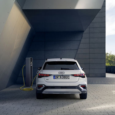 Die Rückseite eines weißen Audi A3 allstreet TFSI e an der Ladesäule vor einer modernen Betonfassade