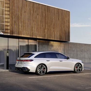 Ein weißer Audi A5 Avant e-hybrid lädt an einer Ladesäule vor einem modernen Haus mit einer Fassade aus Glas, Beton und Holzpanelen.