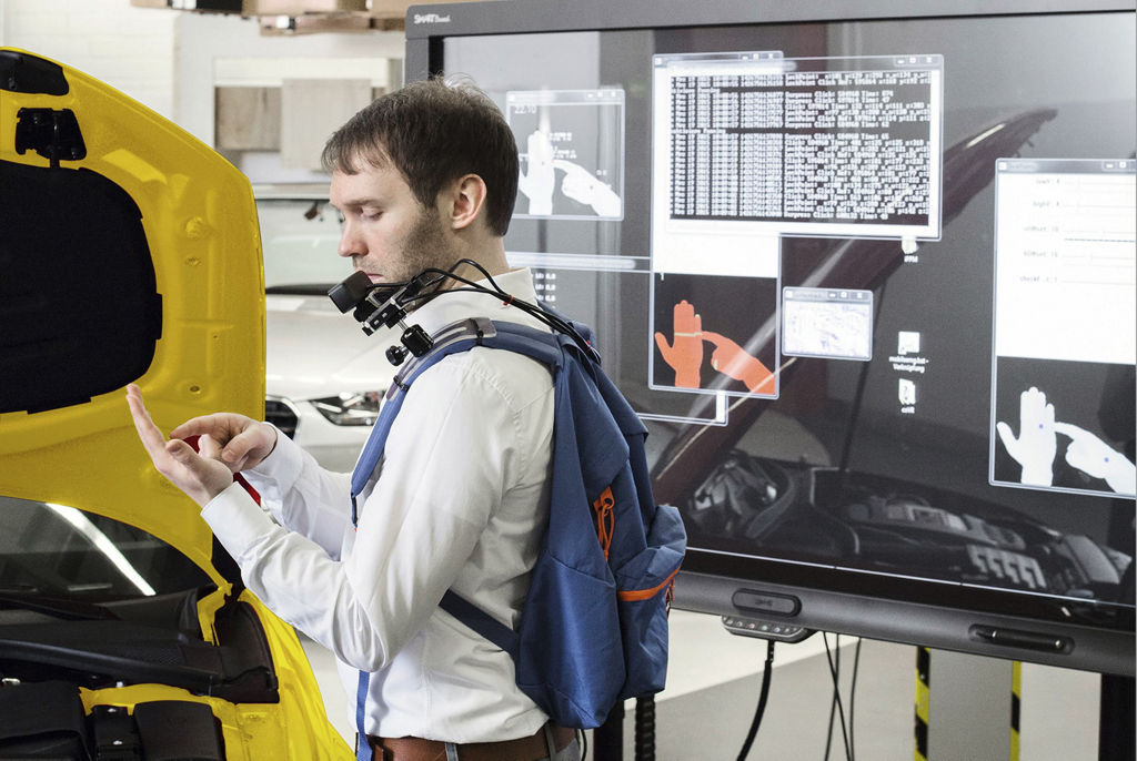 The photo shows an Audi employee wearing a backpack that holds a technical device for hand projection. In testing the projection technology, the technician looks at his hand.