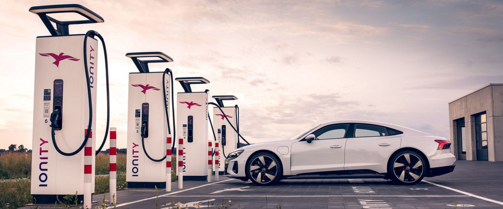 A white Audi e-tron GT quattro stands at a charging station in front of a sunset
