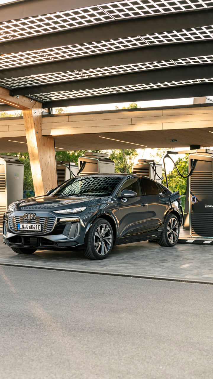An Audi Q6 Sportback e-tron is charging at a charging station at the Audi Charging Hub. The charging station is covered, and the sun is shining through the roof.