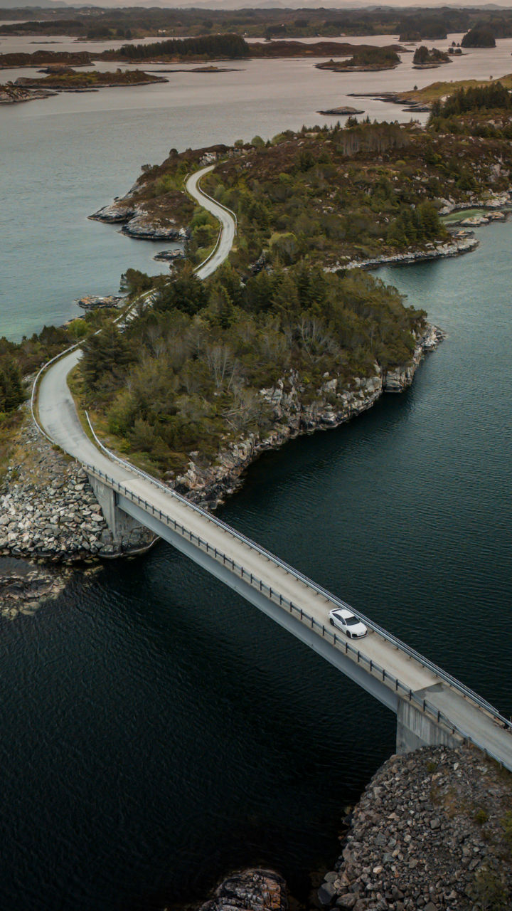 Ein Audi A6 Sportback e-tron in Gletscherweiß Metallic überquert eine Brücke in der zerklüfteten Küstenlandschaft Norwegens. Im Hintergrund befinden sich zahlreiche kleine Inseln. 