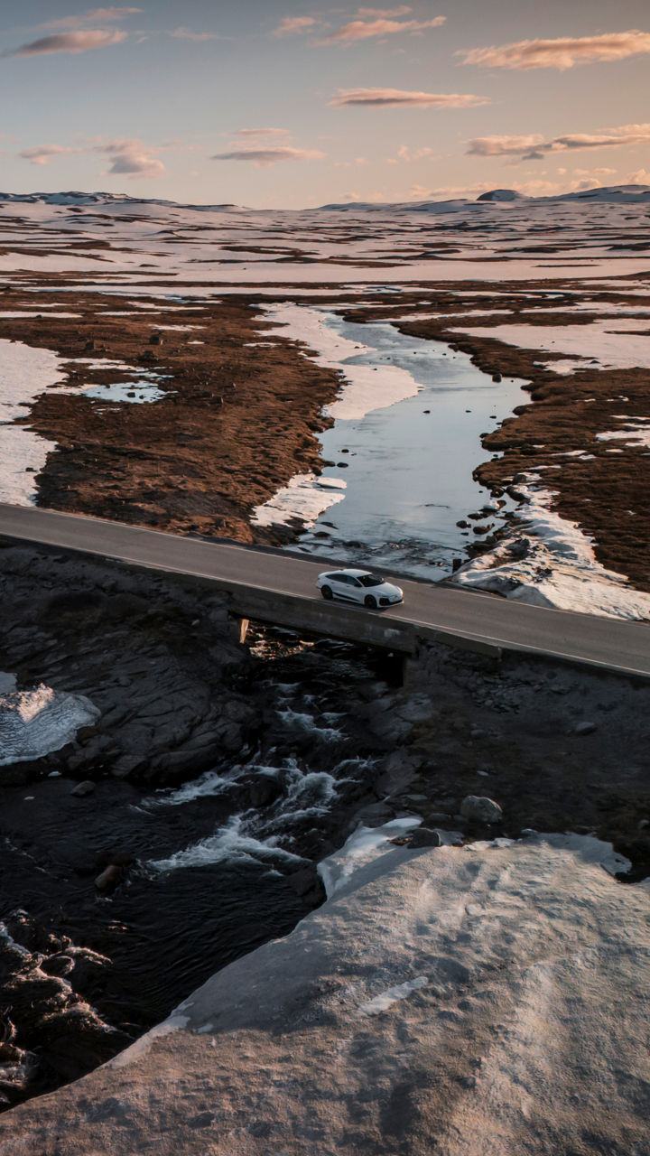 An Audi A6 Sportback e-tron drives along a road through a hilly landscape covered in snow, with the sun setting in the background.