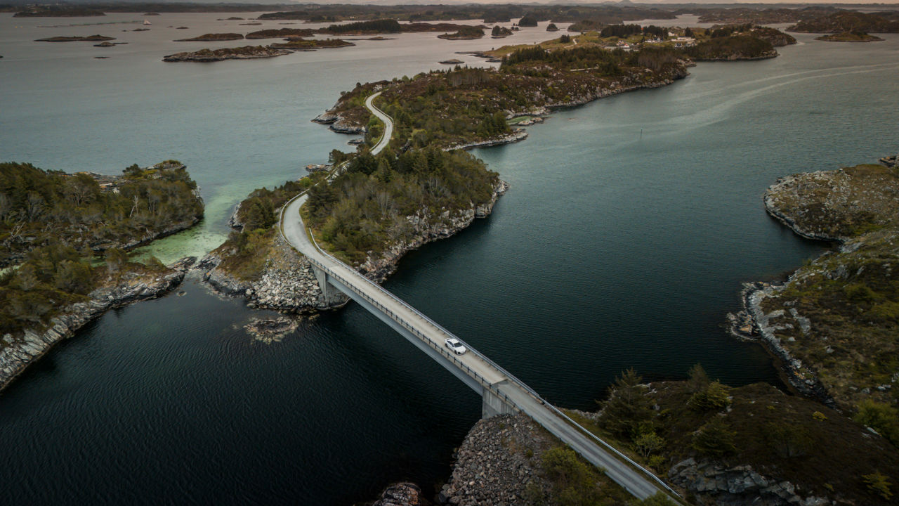 An Audi A6 Sportback e-tron in Glacier White Metallic crosses a bridge in Norway's rugged coastal landscape. Numerous small islands can be seen in the background.