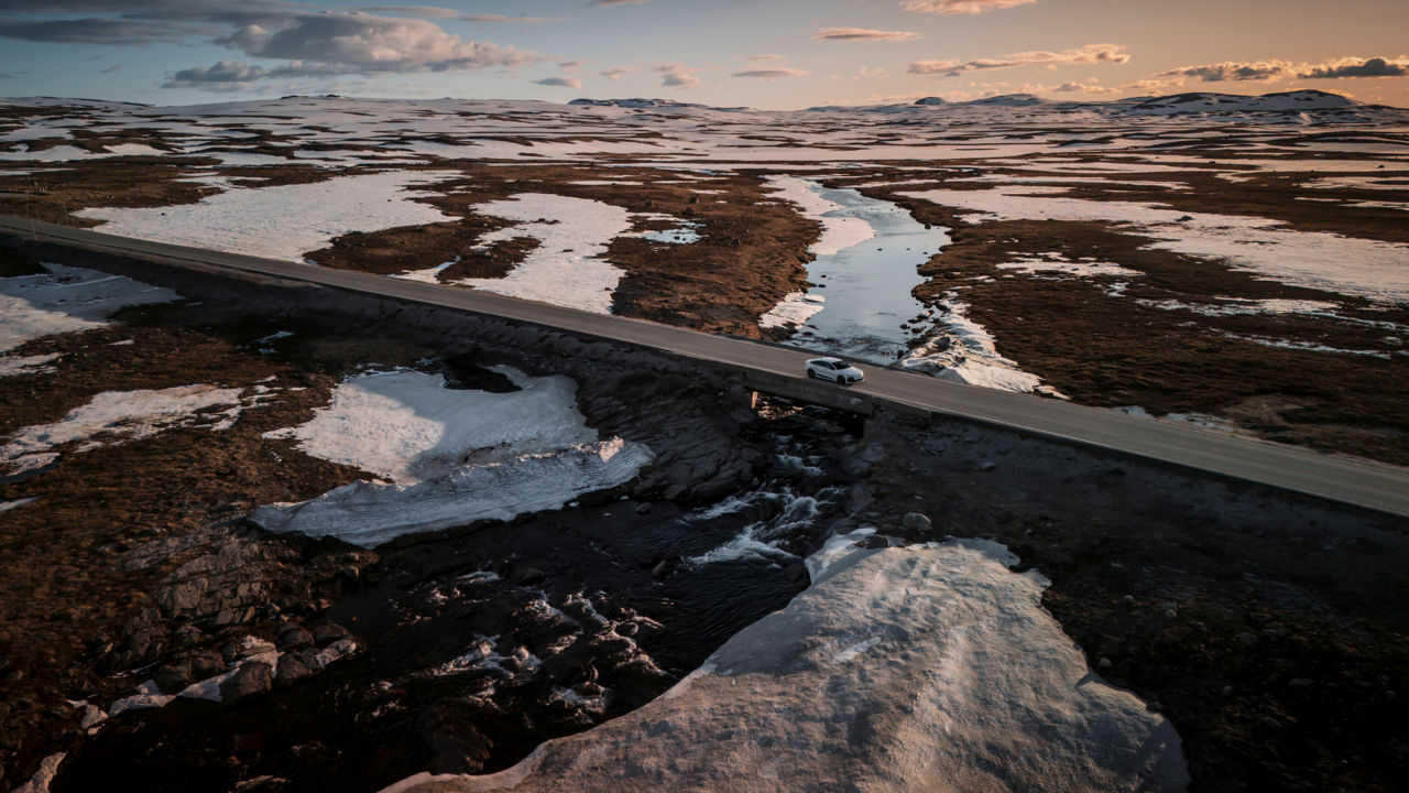 An Audi A6 Sportback e-tron drives along a road through a hilly landscape covered in snow, with the sun setting in the background.