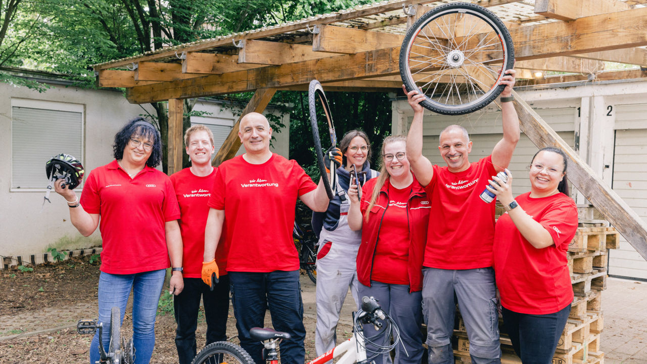 Employees in red T-shirts stand in front of a garage and hold bicycle parts in the air