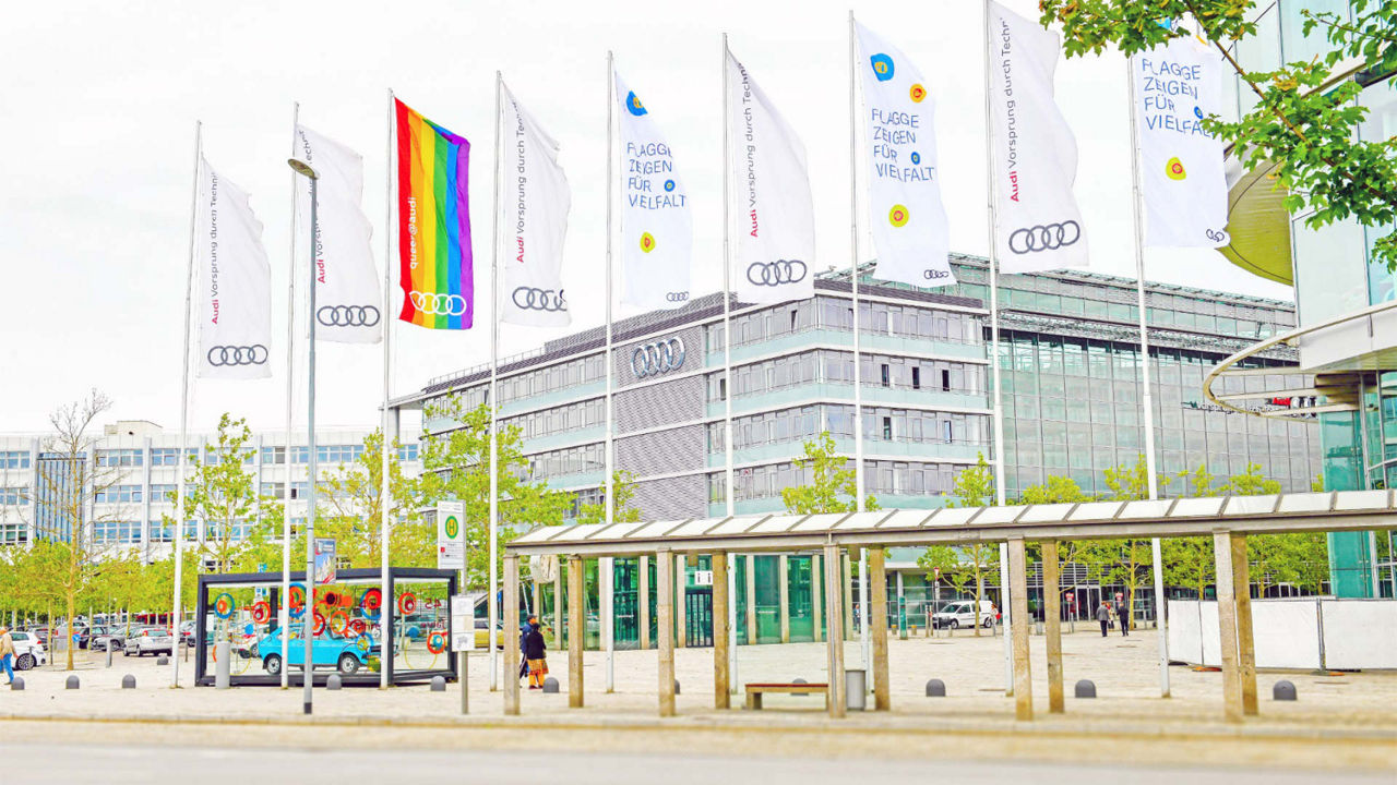 Eine Audi-Gebäudeumgebung mit mehreren Fahnenmasten. Die Fahnen zeigen das Audi-Logo, und eine Fahne zeigt eine Regenbogenflagge als Symbol für Vielfalt und Inklusion.