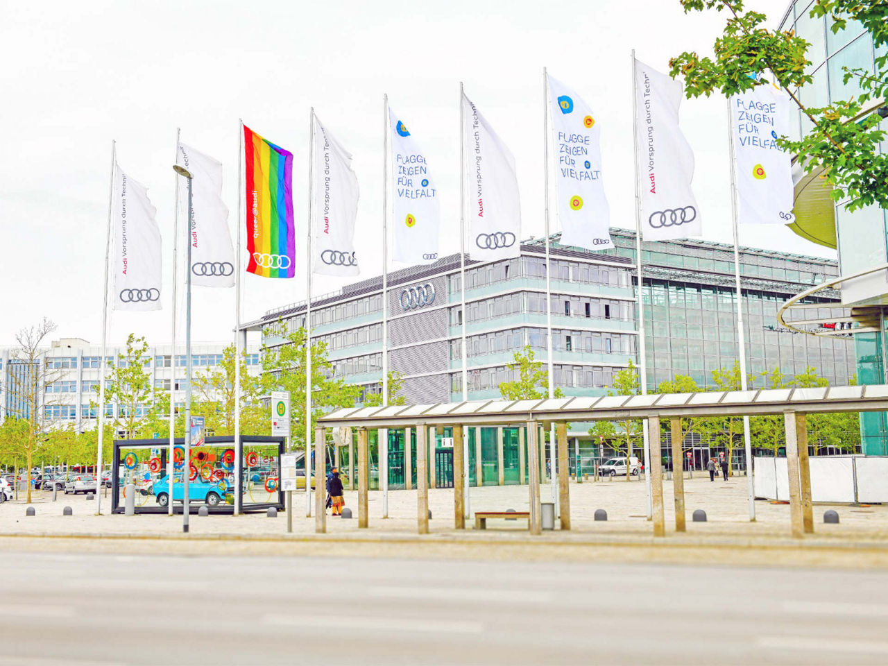 Eine Audi-Gebäudeumgebung mit mehreren Fahnenmasten. Die Fahnen zeigen das Audi-Logo, und eine Fahne zeigt eine Regenbogenflagge als Symbol für Vielfalt und Inklusion.