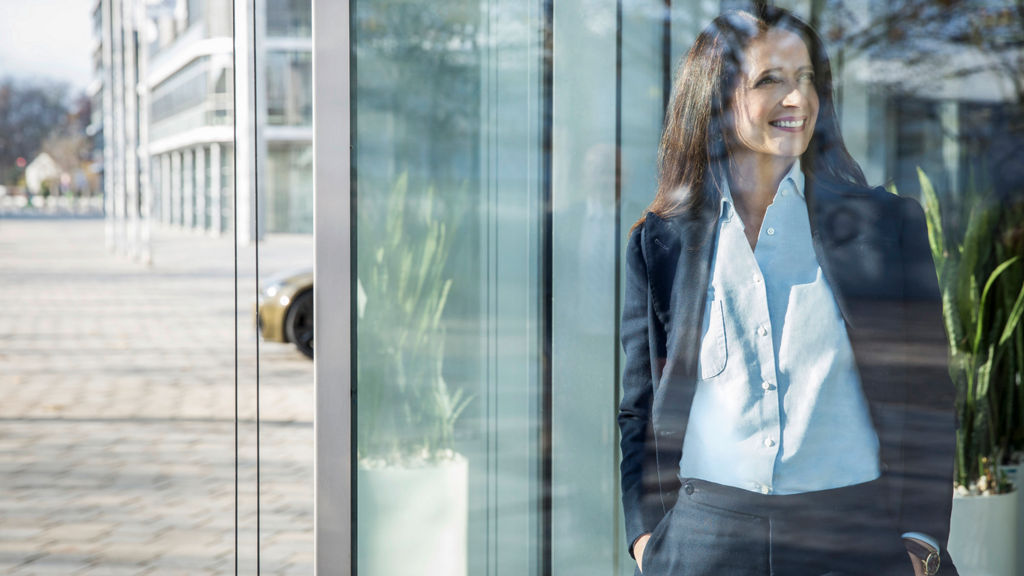 Eine Frau in Business-Kleidung steht lächelnd hinter einer Glaswand und blickt nach draußen. Im Hintergrund sind moderne Gebäude und Pflanzen zu sehen.