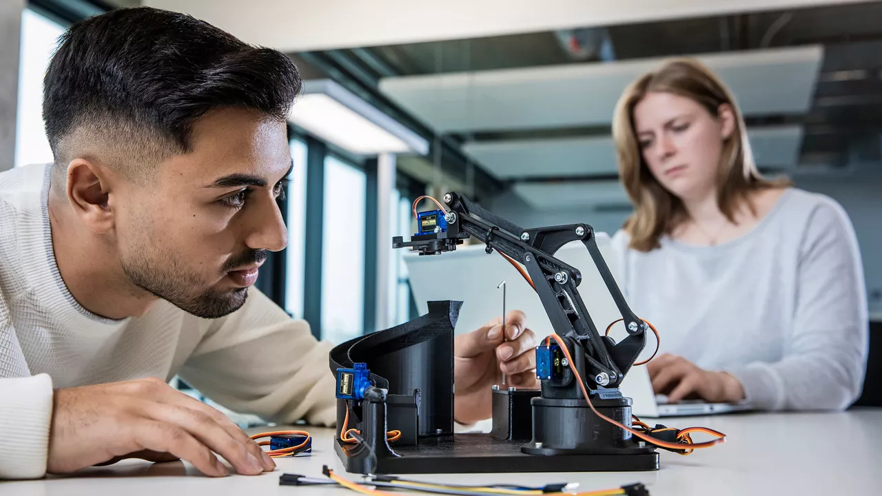 A person in the foreground is working on a small robotic arm. In the background, a person is working on a laptop.