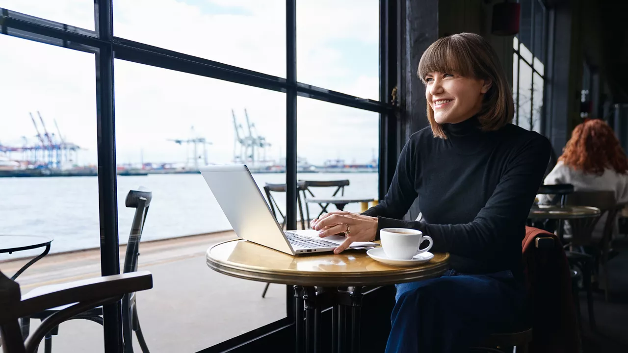 A person sits in front of a laptop in a café. A harbour can be seen in the background.